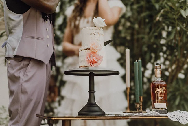 Gâteau de mariage blanc à étages décoré de roses, entouré de bougies et d'une bouteille sur une table élégante.