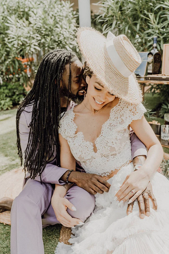 Couple complice en tenue de mariage mauve et dentelle blanche, posant tendrement dans un jardin ensoleillé.