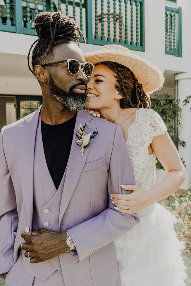 Couple marié complice : homme en costume lilas et femme en robe blanche et chapeau de paille, ambiance romantique et solaire.