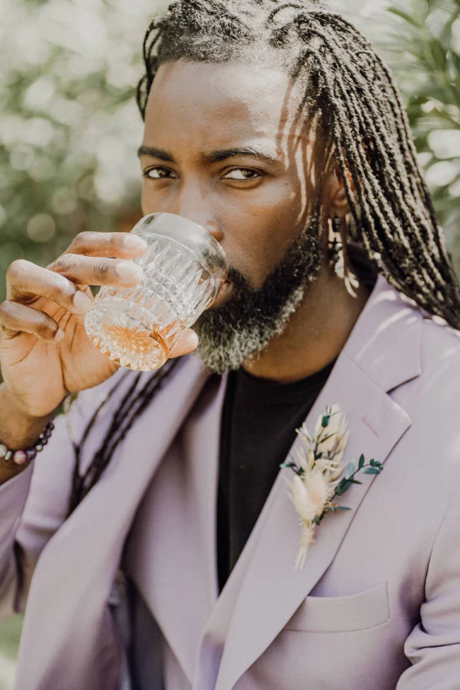 Homme élégant en costume mauve et tresses, buvant dans un verre ciselé dans une ambiance lumineuse et romantique.
