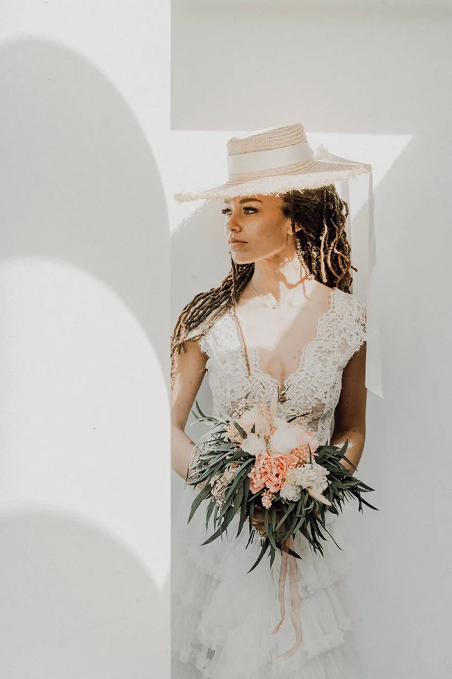Mariée bohème en robe de dentelle blanche et chapeau paille, tenant un bouquet floral aux tons poudrés et verts.