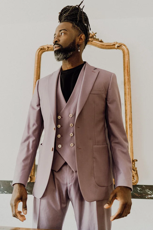 Homme noir aux dreadlocks portant un costume trois-pièces mauve poudré devant un miroir doré.