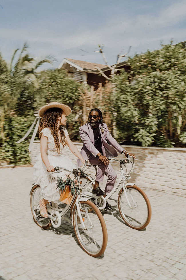 Jeunes mariés élégants à vélo dans un jardin, ambiance romantique et lumineuse aux tons pastel.