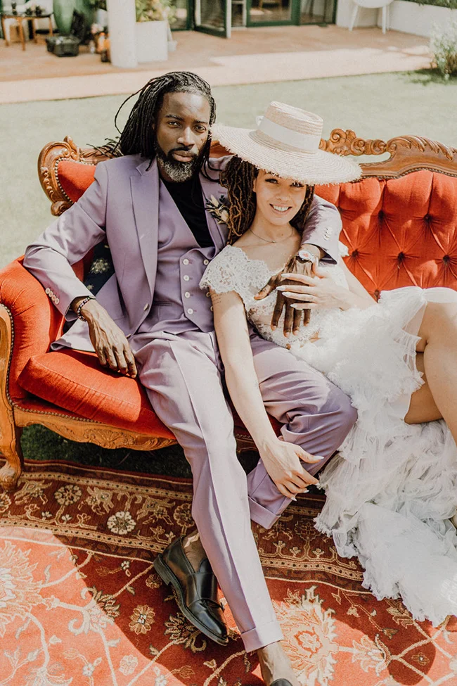Couple élégant posant sur un canapé rouge : il porte un costume lilas, elle une robe blanche et un chapeau de paille.