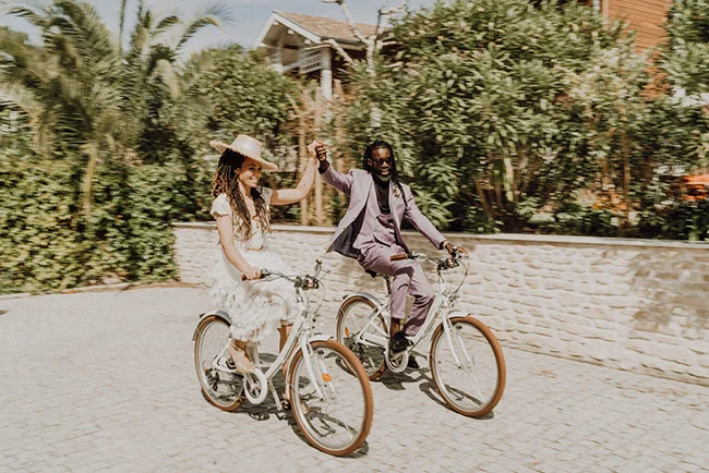 Jeunes mariés élégants se tenant la main à vélo dans un jardin luxuriant, ambiance romantique et lumineuse.