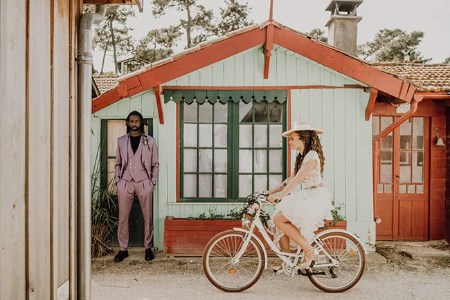 Couple marié stylé devant un chalet bleu pastel, la mariée à vélo et le marié en costume mauve, ambiance romantique.