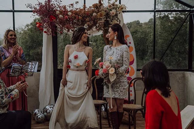 Deux mariées souriantes en robes blanches sous une arche fleurie, dans une ambiance festive et rétro.