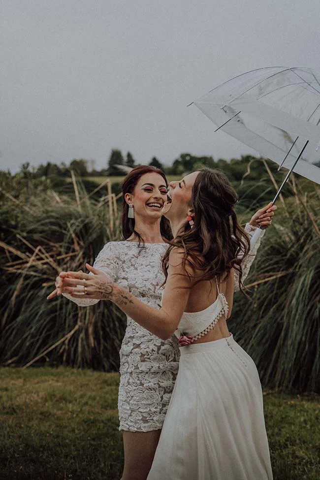 Deux mariées rient sous un parapluie transparent, portant des robes blanches en dentelle et satin dans un décor naturel.