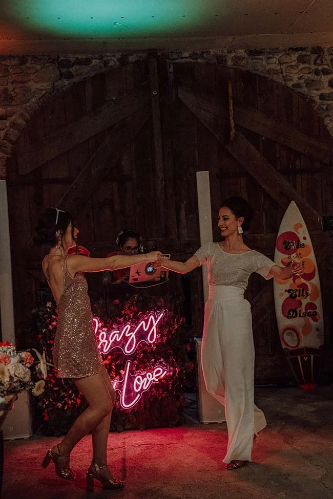 Deux femmes dansent joyeusement devant un néon rose Crazy Love lors d'une soirée mariage disco rétro.