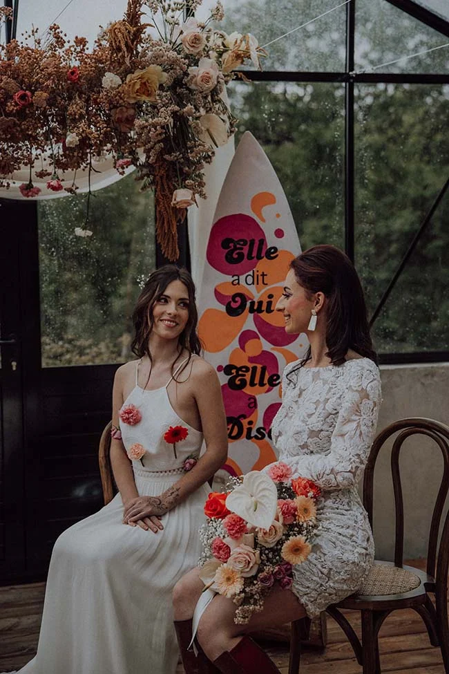Deux mariées en robes blanches posant devant un surf rétro coloré et des fleurs séchées dans une ambiance disco.