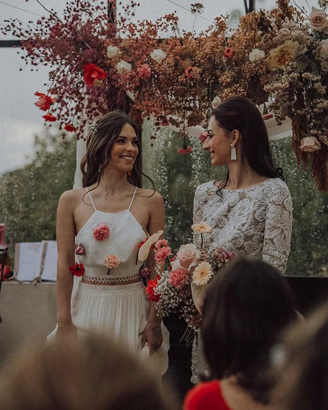 Deux femmes souriantes en robes blanches sous une arche florale aux tons automnaux, ambiance romantique et rétro.