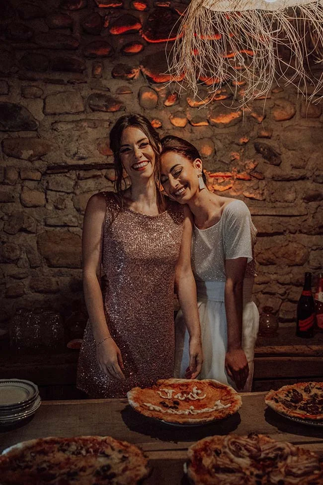 Deux femmes souriantes en robes de soirée posent devant des pizzas dans un cadre rustique et chaleureux.