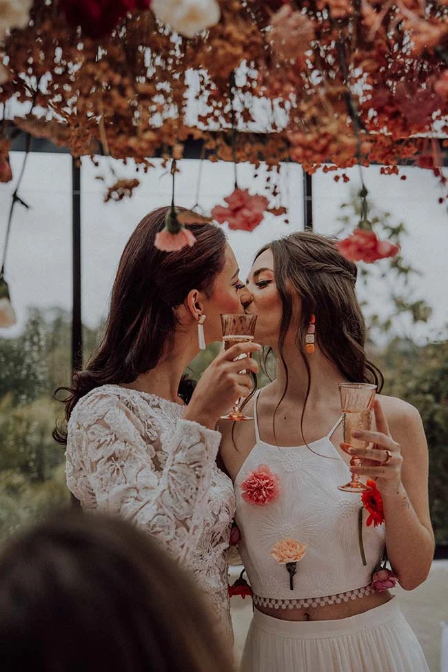 Deux mariées s'embrassent avec des coupes de champagne sous une pluie de fleurs séchées aux tons chauds.