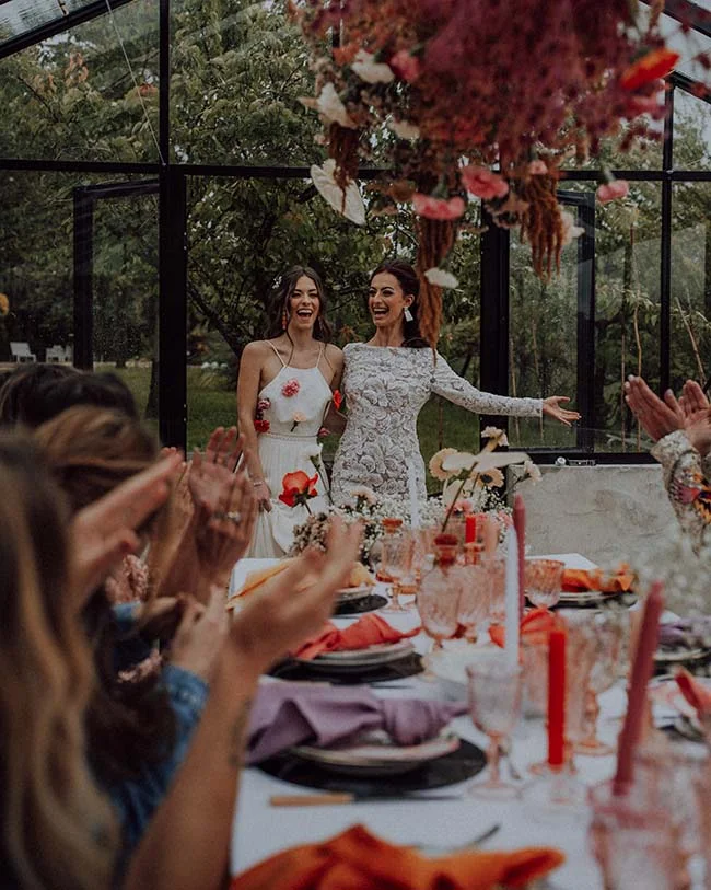 Deux mariées rayonnantes et riantes devant leurs invités lors d'un dîner festif sous une verrière fleurie.