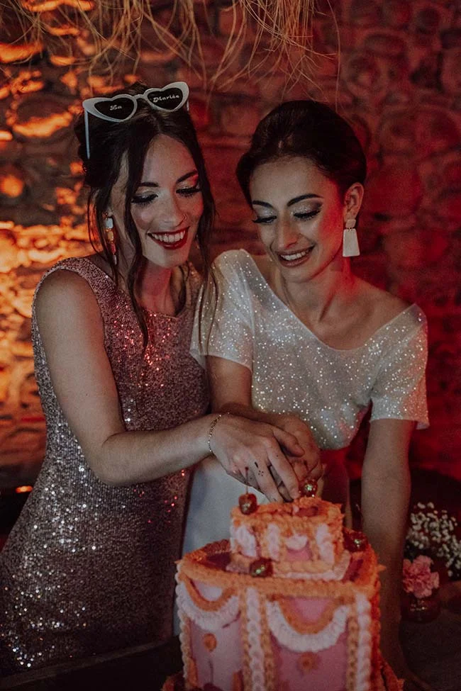 Deux femmes souriantes en robes pailletées découpent un gâteau rose dans une ambiance festive et chaleureuse.