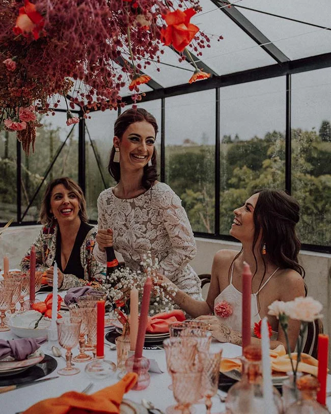 Trois femmes souriantes à une table festive et colorée sous une verrière, décorée de fleurs suspendues et de bougies.
