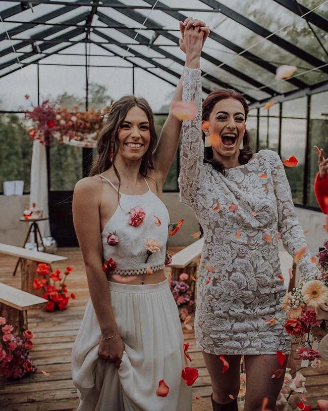 Deux mariées joyeuses en robes blanches se tiennent la main sous une pluie de pétales dans une serre lumineuse.