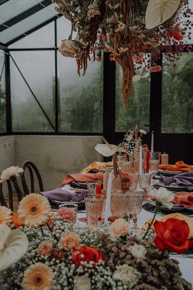 Table de mariage rétro aux tons terracotta et poudrés, sous une installation florale suspendue dans une verrière.