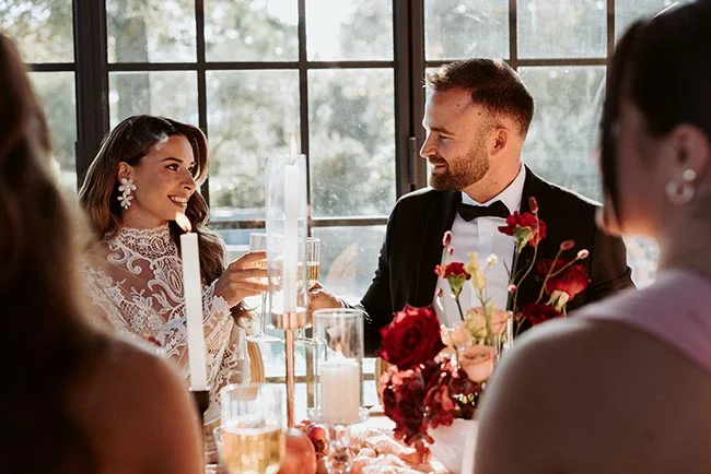 Jeunes mariés élégants s'échangeant un regard complice lors d'un dîner romantique aux chandelles et roses rouges.