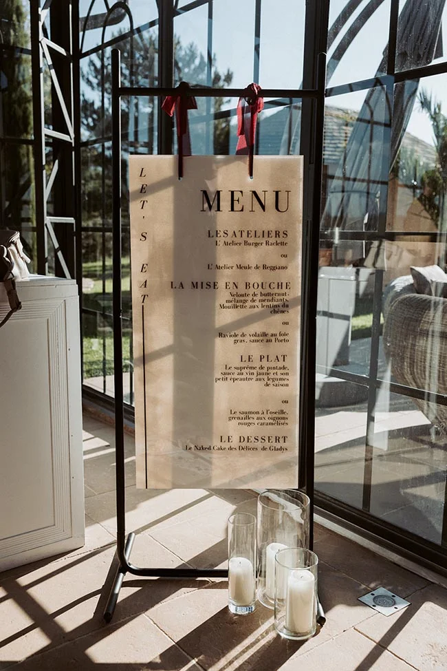 Menu de mariage élégant sur chevalet noir, tons crème et rouge, installé dans une verrière lumineuse.