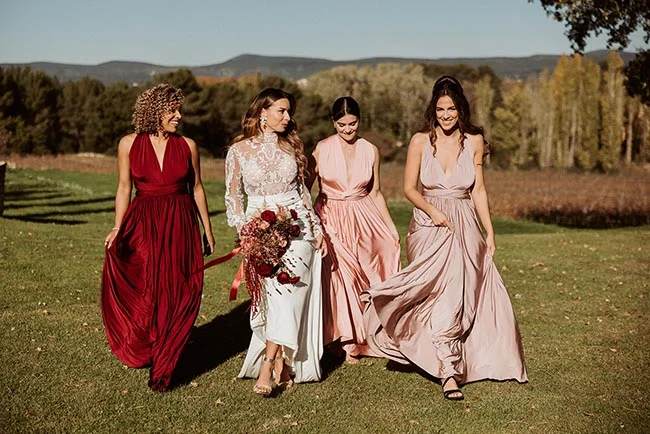 Une mariée en robe blanche et trois demoiselles d'honneur en robes rouges et roses marchent dans un paysage provençal.