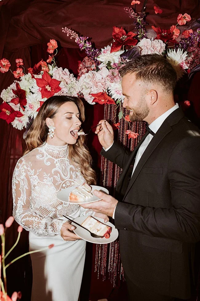 Jeunes mariés partageant une part de gâteau devant un décor floral rouge et blanc dans une ambiance romantique.