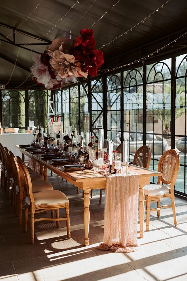 Table de mariage élégante en bois avec chemin de table rose et décorations rouges sous une verrière lumineuse.