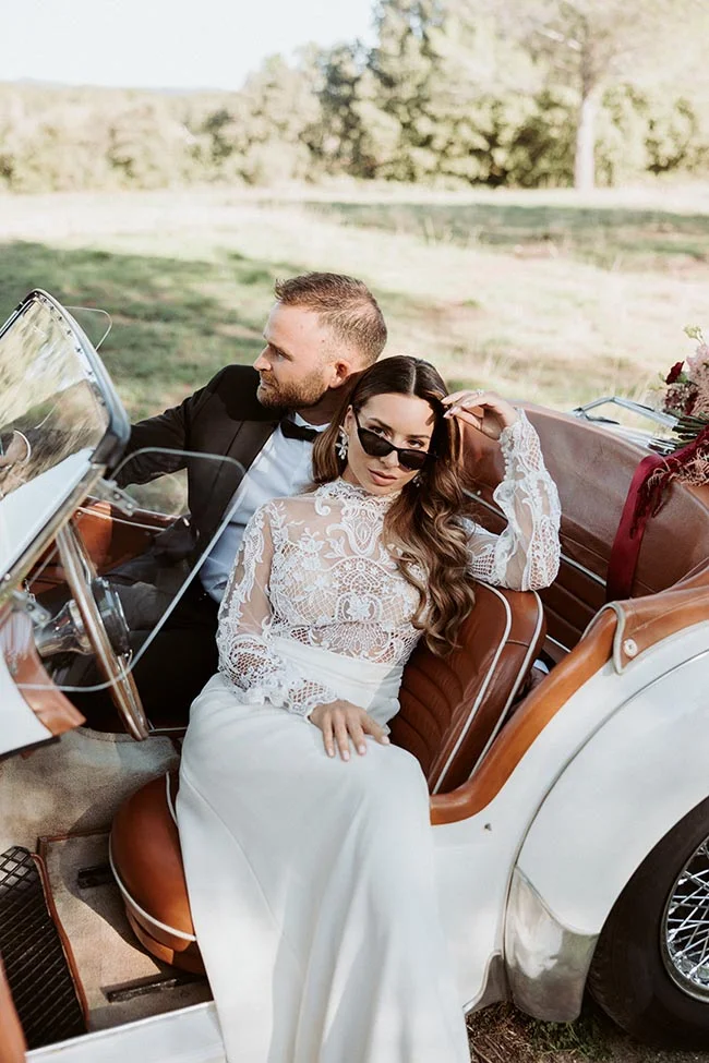 Couple élégant dans une voiture ancienne blanche, la mariée en robe de dentelle et le marié en smoking, ambiance lumineuse.