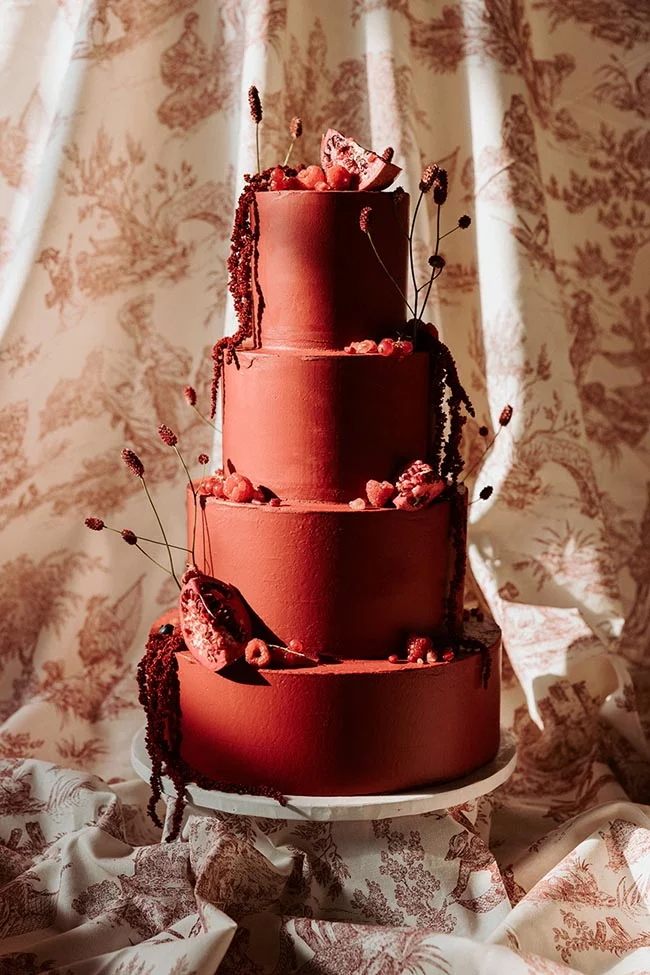 Gâteau de mariage à quatre étages rouge terracotta, décoré de baies et fleurs séchées sur fond de tissu floral.