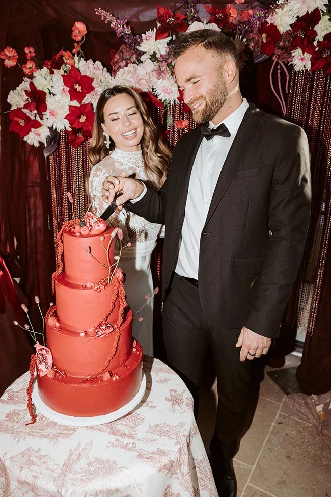 Un couple élégant découpe un gâteau de mariage rouge à trois étages devant un décor floral rouge et blanc.
