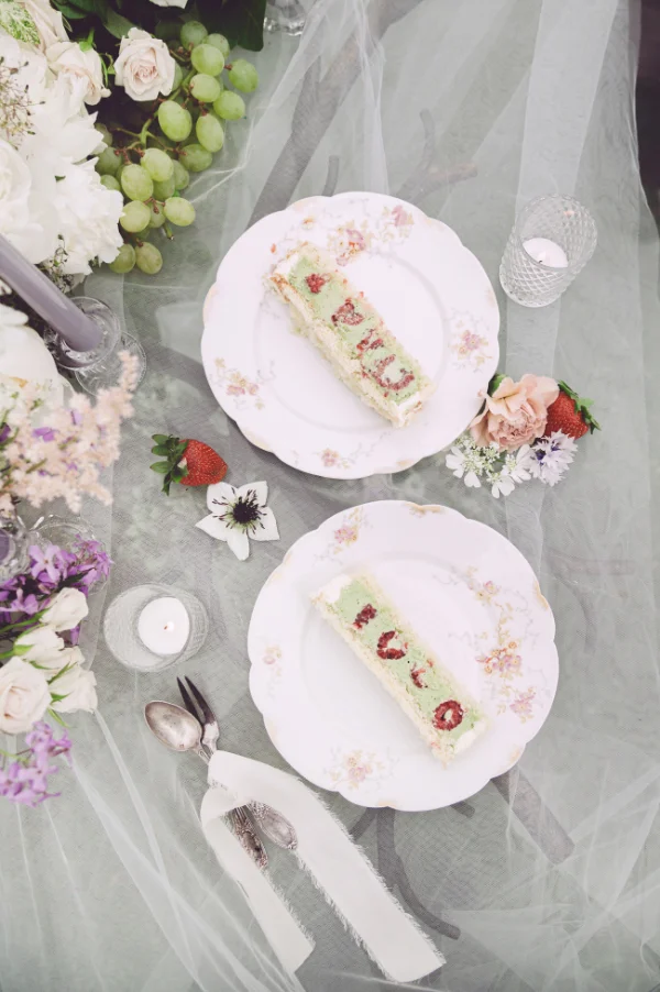 Deux parts de gâteau aux fruits sur des assiettes vintage, entourées de fleurs et de raisins sur un nappage vaporeux.