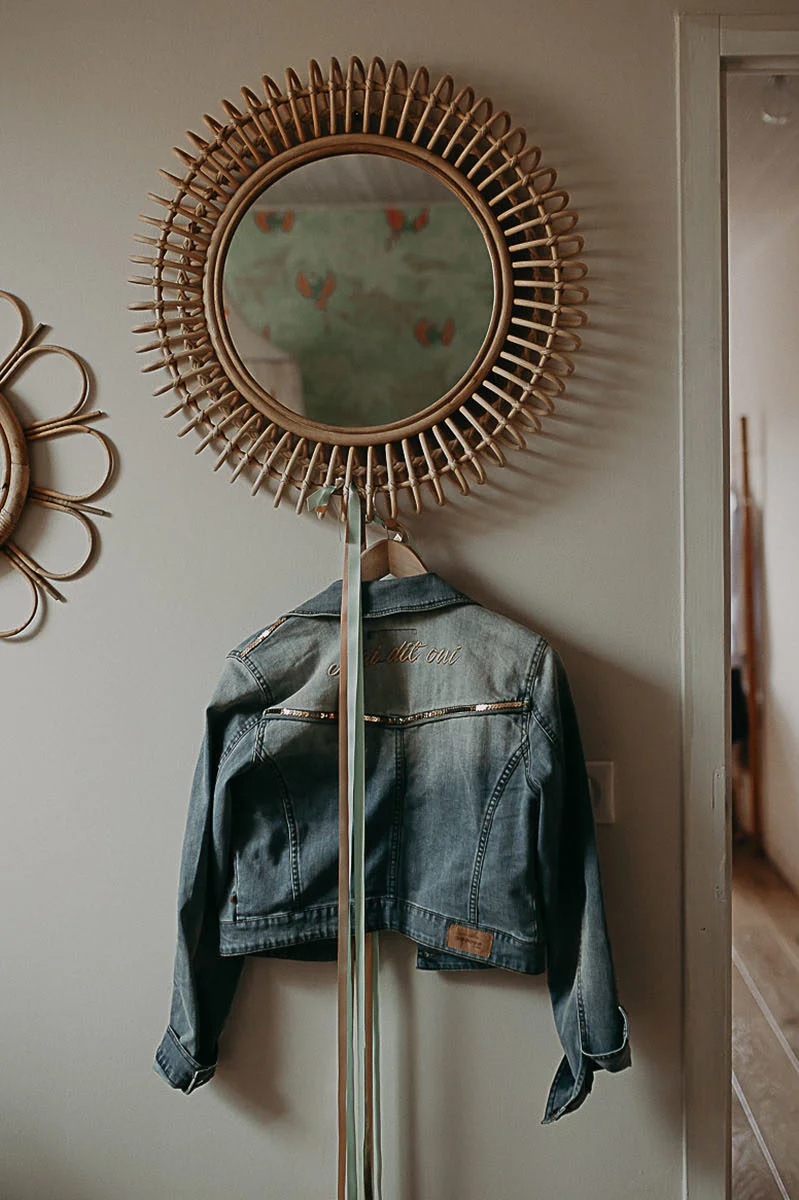 Veste en jean personnalisée suspendue sous un miroir soleil en rotin dans un intérieur aux tons neutres et doux.