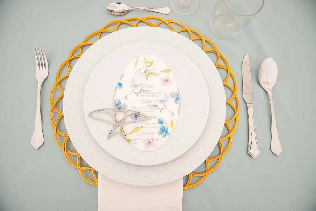 Dressage de table romantique avec assiettes blanches, set de table jaune et menu floral sur nappe bleu pâle.
