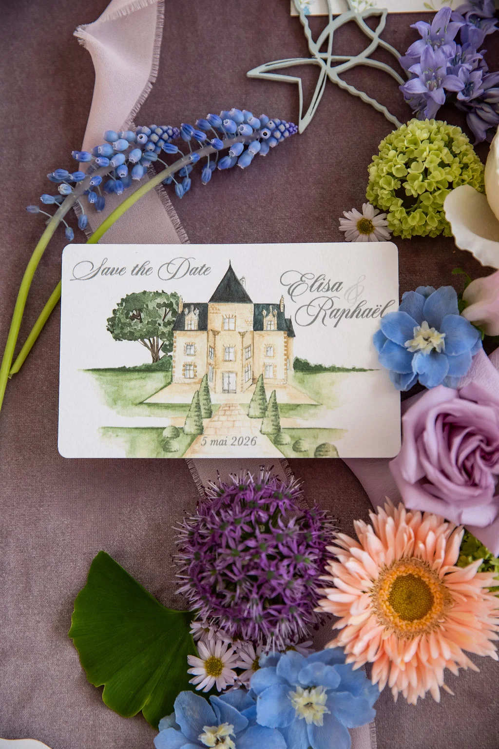 Carte Save the Date illustrée d'un château, entourée de fleurs pastel et de rubans sur un fond mauve romantique.