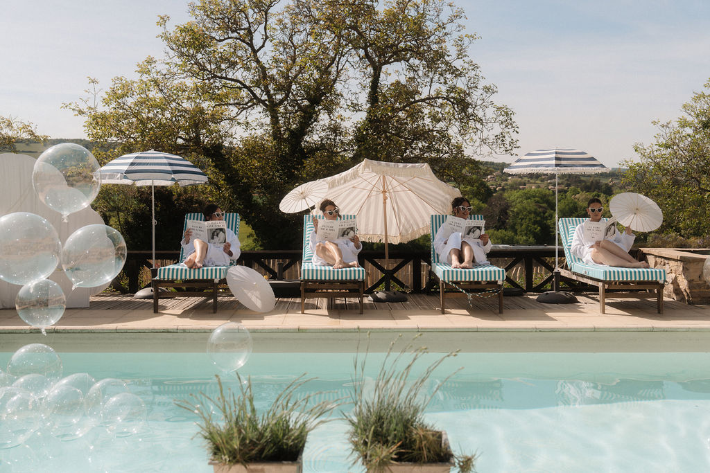 4 femmes sur un transat rayé bleu et blanc abrité par des omrelles blanches au bord d'une piscine