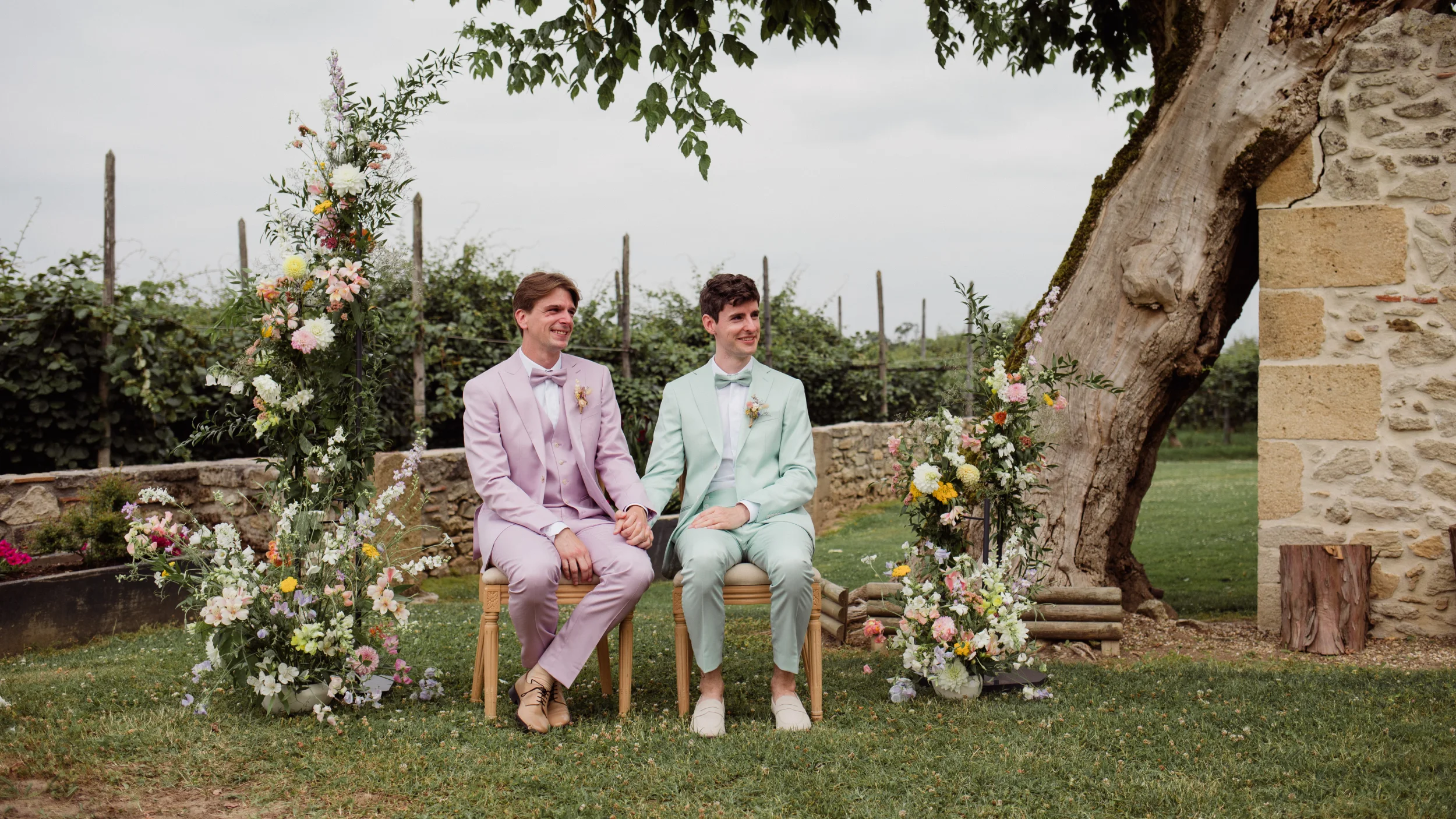 Deux hommes en costumes pastel rose et vert menthe assis côte à côte dans un jardin fleuri et romantique.