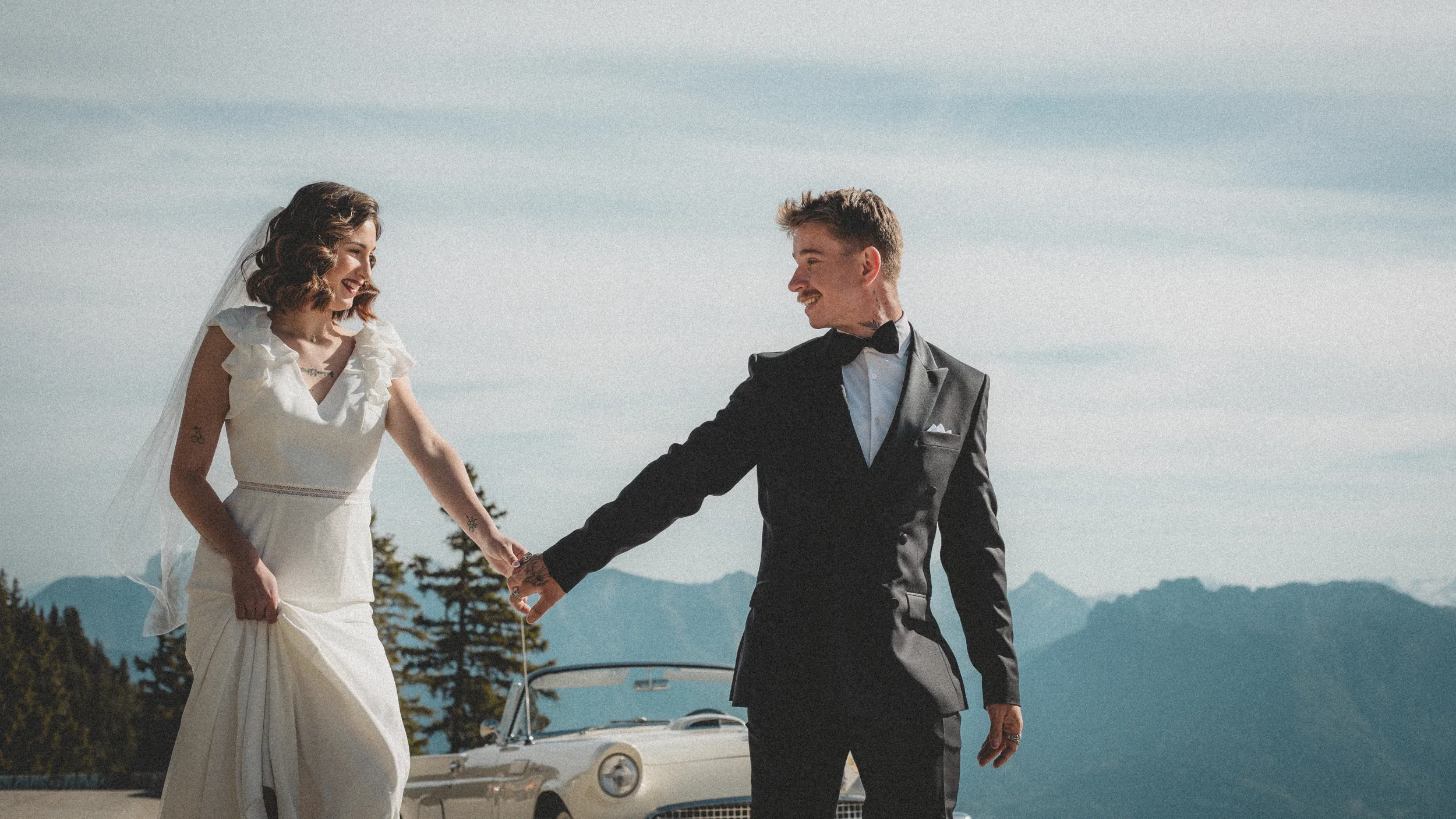 Une mariée et un mari en costume sombre tenant la main, visant un paysage montagneux avec un vintage automobile en