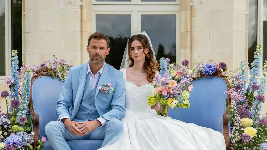 Couple de mariés élégants assis sur un canapé bleu entouré de fleurs colorées devant un domaine luxueux.