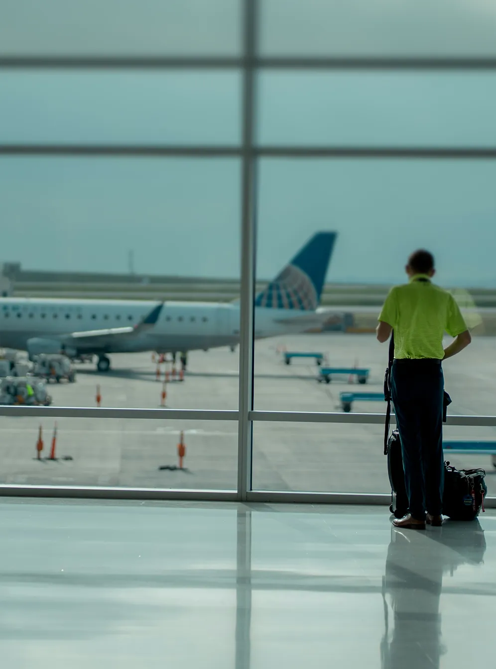 Person in grünem Hemd steht mit Gepäck vor einem großen Fenster am Flughafen, durch das ein Flugzeug sichtbar ist.