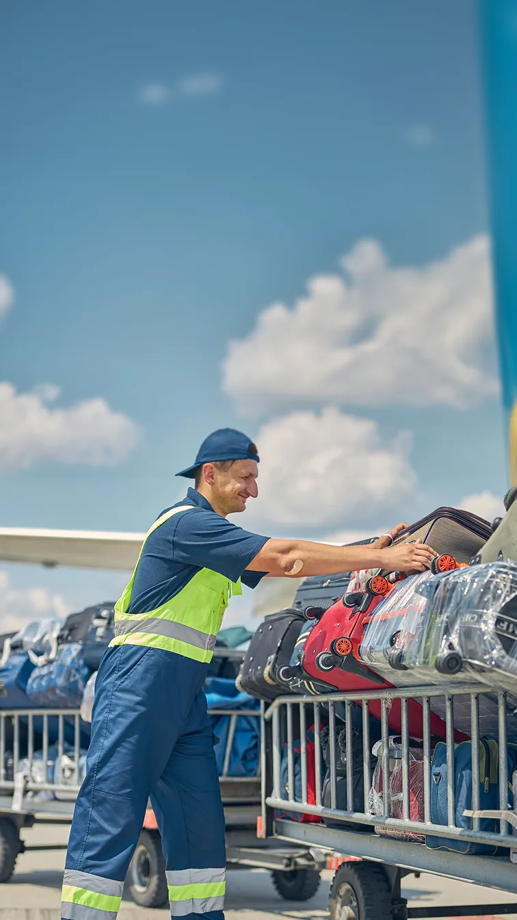 Flughafenmitarbeiter sortiert Koffer auf Gepäckwagen bei klarem Himmel.