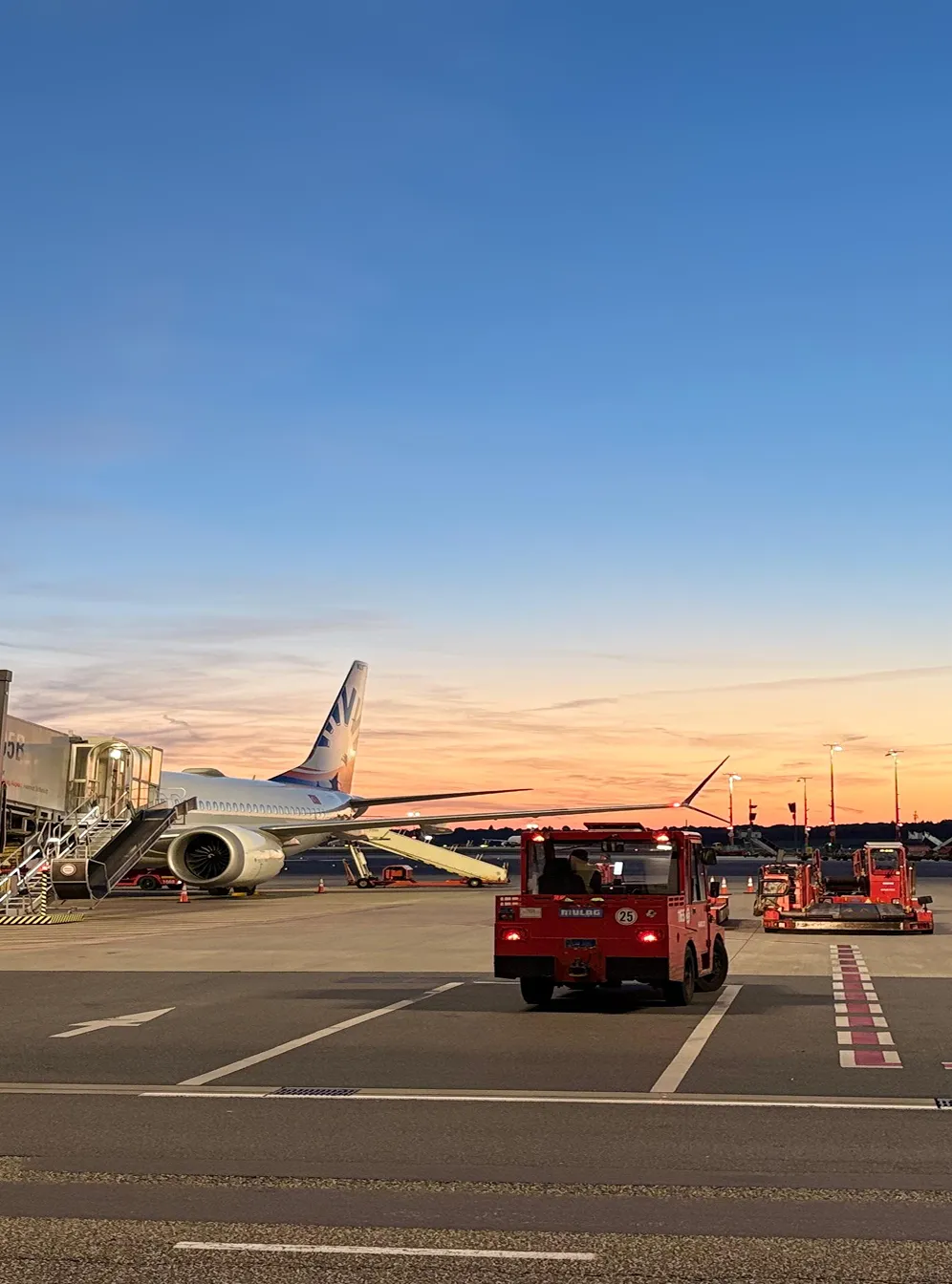 Flugzeug mit angedocktem Passagiersteig und mehreren roten Flughafenfahrzeugen bei Sonnenuntergang auf dem Rollfeld.