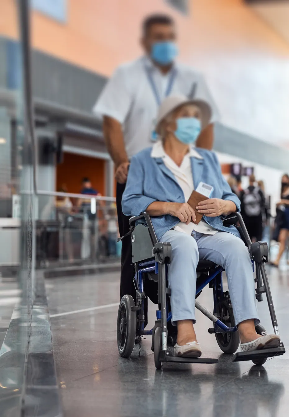 Ältere Frau mit Mundschutz im Rollstuhl wird von einem Helfer in einem Flughafen geschoben.