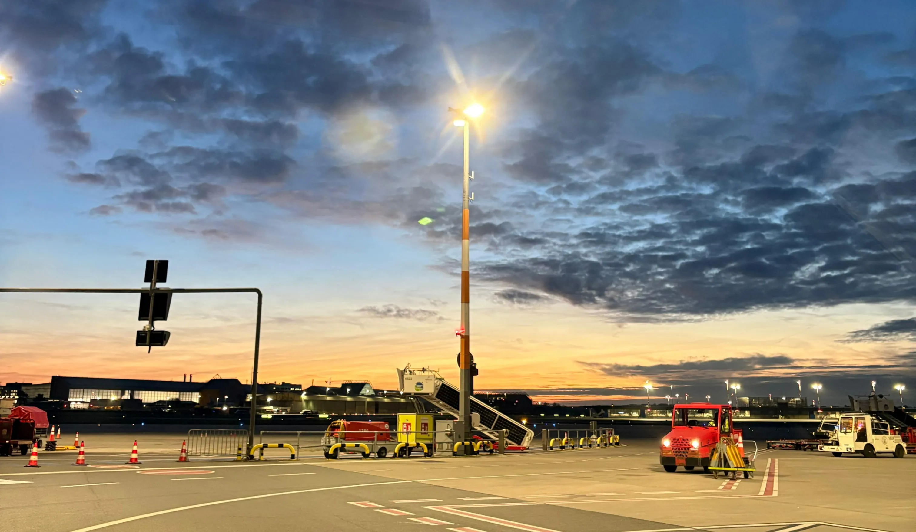Flughafenvorfeld bei Sonnenuntergang mit Fahrzeugen und beleuchteten Gebäuden im Hintergrund.