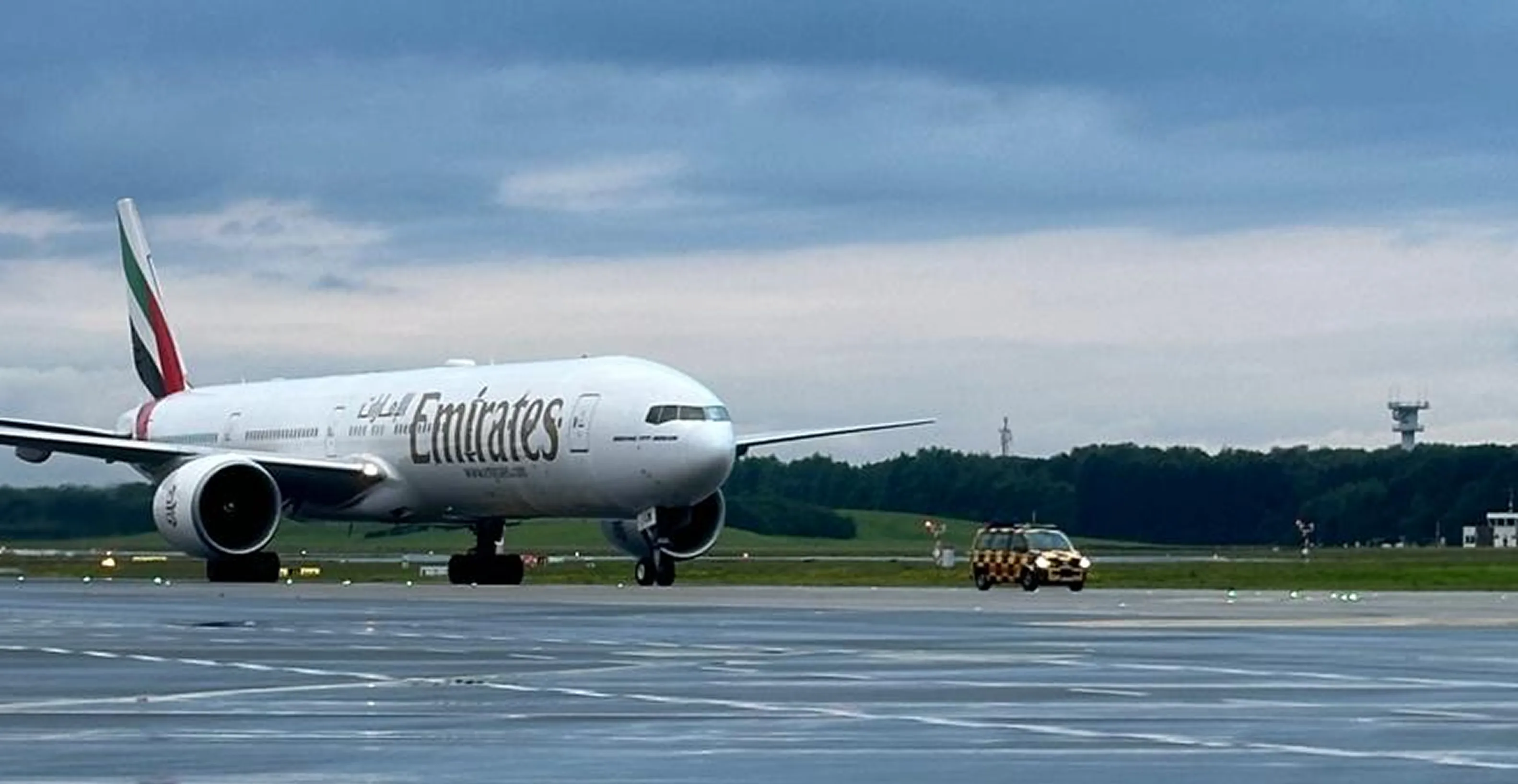 Ein Emirates-Flugzeug rollt auf der nassen Start- und Landebahn eines Flughafens bei bewölktem Himmel, begleitet von einem gelb-schwarzen Fahrzeug.
