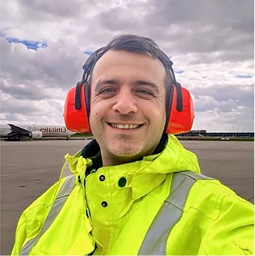 Lächelnder Mann in gelber Sicherheitsjacke und roten Gehörschützern auf einem Flughafen rollfeld.