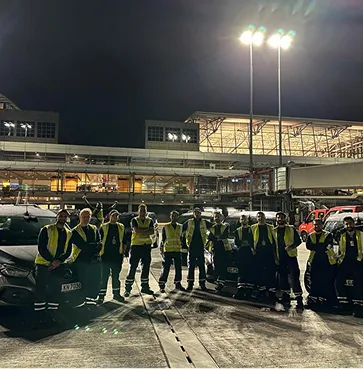 Gruppe von Sicherheits- oder Flughafenmitarbeitern in gelben Warnwesten stehen nachts auf einem Parkplatz vor einem beleuchteten Flughafengebäude.