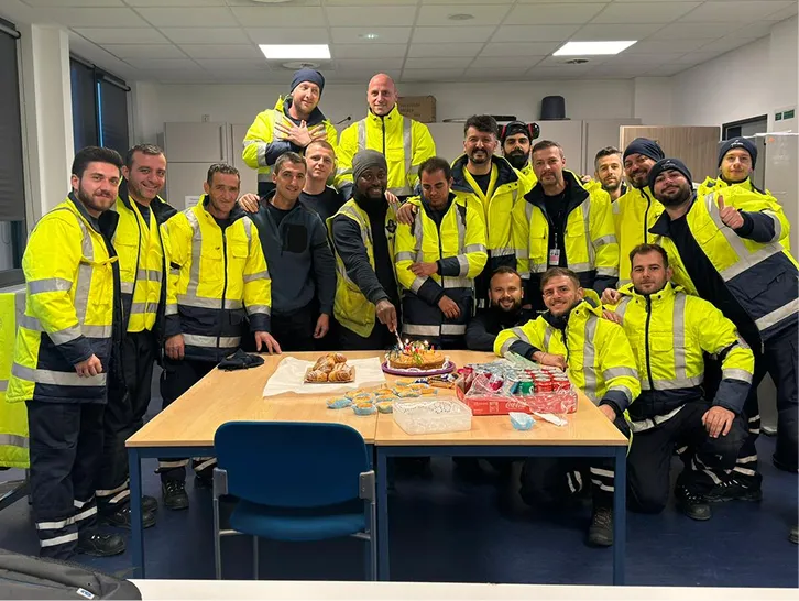 Gruppe von Arbeitern in gelben Sicherheitsjacken, die um einen Tisch mit Kuchen und Snacks stehen und lächeln.