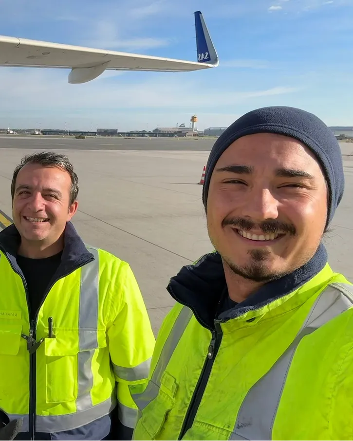 Zwei Männer in leuchtend gelben Sicherheitsjacken lächeln auf einem Flughafenrollfeld unter einem Flugzeugflügel.