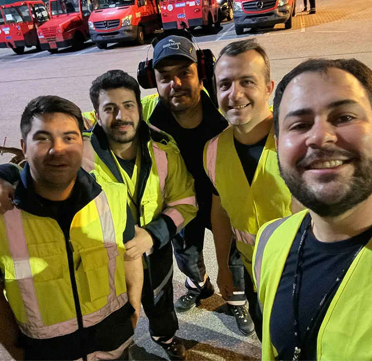 Fünf Männer in gelben Sicherheitswesten posieren vor dem Hintergrund mehrerer roter Fahrzeuge auf einem Parkplatz.