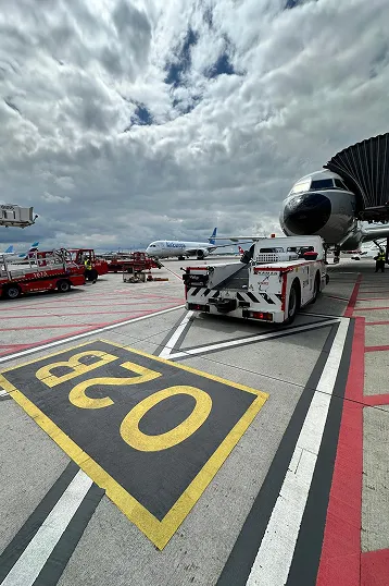 Flugzeug am Gate mit Angriffsbrücke und einem Flugzeugschlepper auf dem Flughafenvorfeld unter bewölktem Himmel.